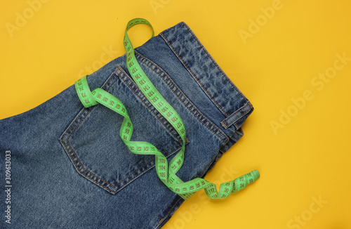 Blue Jeans with measuring tape on yellow background. Concept of weight loss, diet, detox. Top view