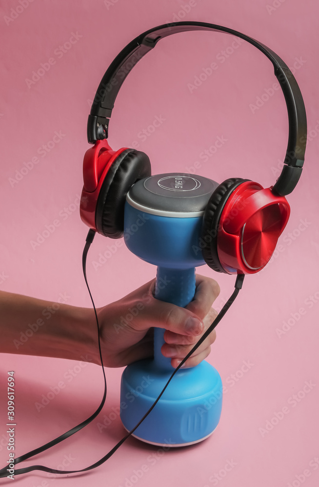 Minimalistic sport and fitness concept. Female hand holding wired headphones and a dumbbell on pink background