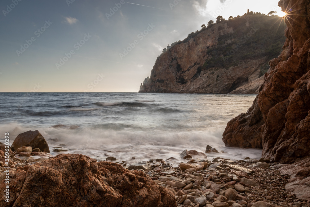 Small rocky beach bathed in the last sun rays with waves breaking on the shore in Cala Giveroleta, Catalonia, Spain.