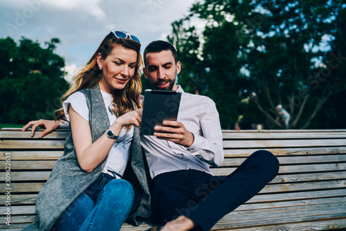 Young positive male and female best friends searching interesting video in webpages on digital tablet connected to wireless internet.Happy couple recreating together at urban setting with gadget