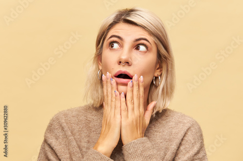 Surprise and astonishment concept. Picture of beautiful young woman with blonde bob hairdo opening mouth widely and widening eyes, being shocked with unexpected news, holding hands on her cheeks