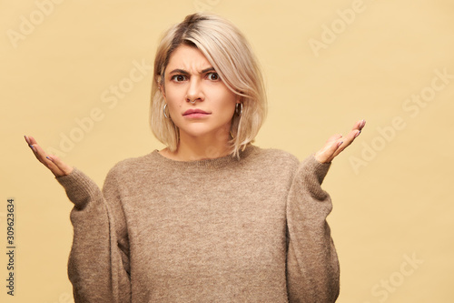 Angry perplexed young blonde woman in beige sweater frowning having indignated look, shrugging shoulders trying to figure out what happened, gesturing emotionally. Blaming, warning, accusing concept photo