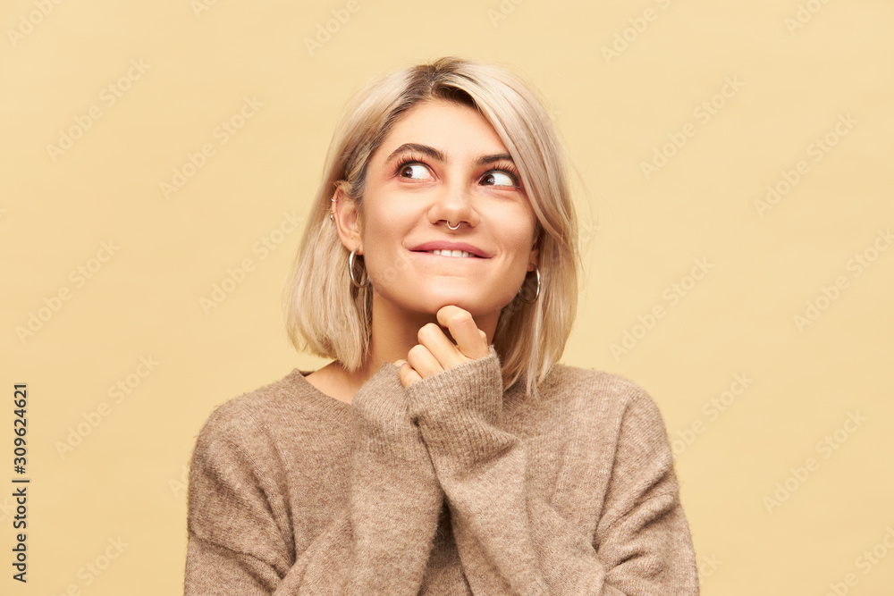 Close up image of charming young blonde female wearing nose ring and bob haircut holding hands under her chin and looking away with playful mysterious smile, playing pranks and doing mischief Close up image of charming young blonde female wearing nose ring and bob haircut holding hands under her chin and looking away with playful mysterious smile, playing pranks and doing mischief