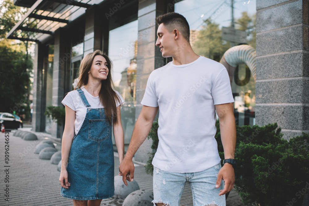 Positive male and female hipster guys holding each other on hands and walking around urban setting in city area,happy couple in love enjoying free time and summer vacations for flirting and recreating