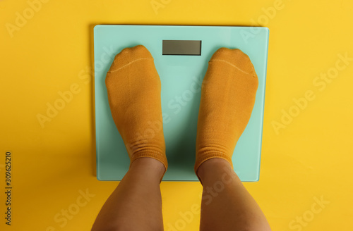 Female legs in socks are standing on the floor scales against yellow background. Top view photo