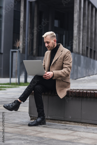 Stylish man sitting on the streets of the city near modern office centre and working with his laptop.