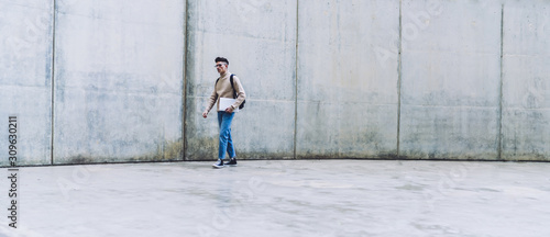 Young male in glasses carrying laptop near grey concrete wall