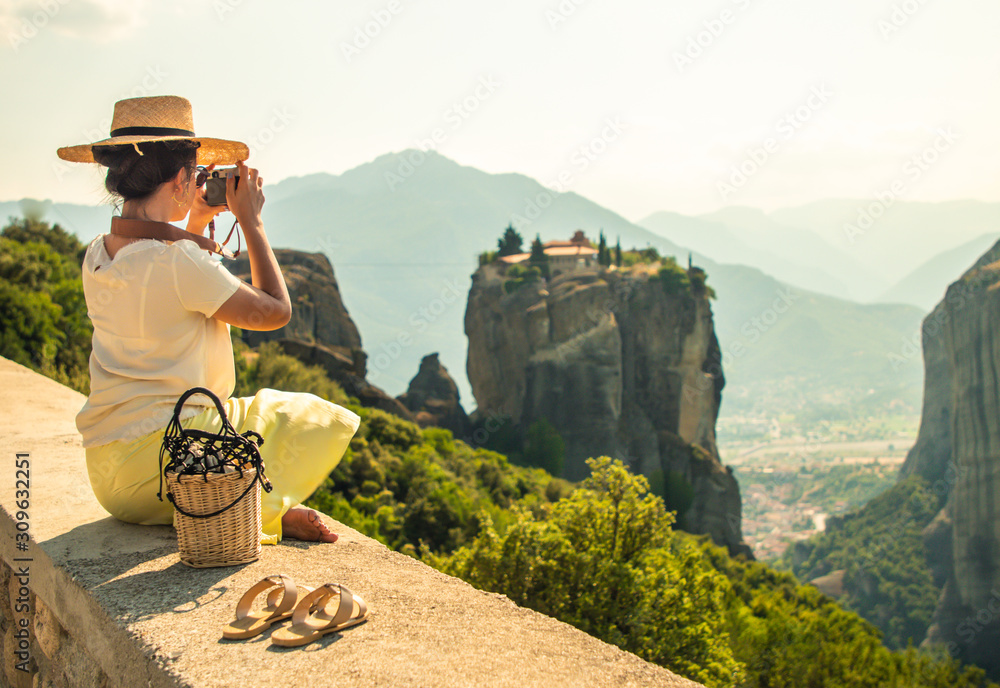 Naklejka premium Young woman with white dress and large hat sitting in front of greece meteor mountains, monastery and village in the background