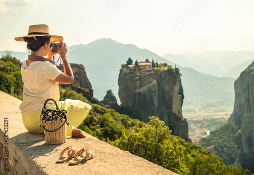 Young woman with white dress and large hat sitting in front of greece meteor mountains, monastery and village in the background photo