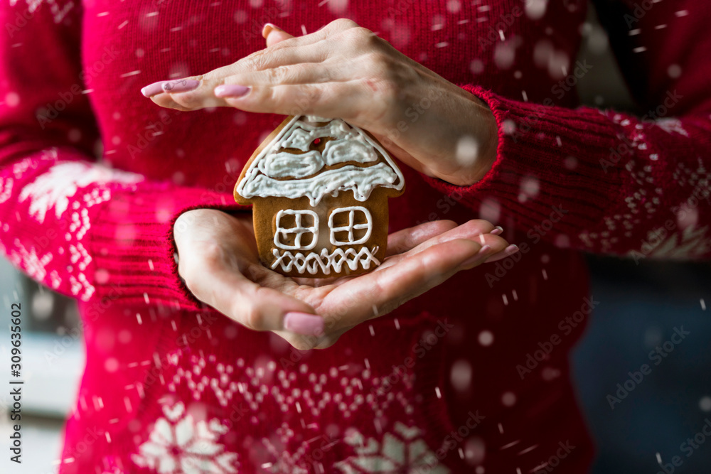 Female hands hold a Christmas painted gingerbread in the form of a house. The model is wearing a red winter sweater with a white Scandinavian pattern. Added falling snow effect.