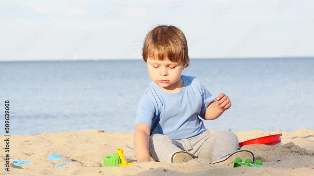 Little child boy sitting on sand, playing with toys on beach seashore. Slow motion Little child boy sitting on sand, playing with toys on beach seashore. Slow motion