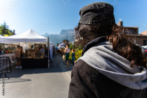 A man wearing a black jacket and baseball cap is seen from the back at a street fair during summer, blurry stalls are in background with copy space
