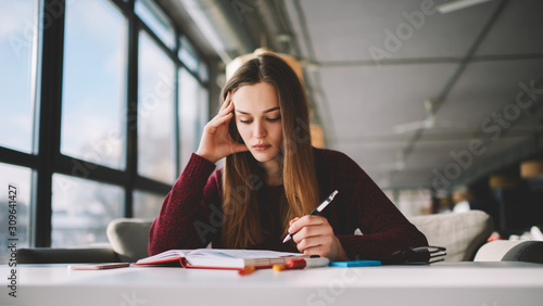 Thoughtful hipster girl studying at school cafeteria while reading literature and writing main theses in notebook, pensive skilled young female student learning concentrated on course work indoors