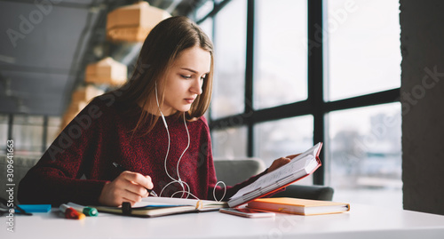 Pensive hipster girl learning language online via earphones using application while writing new words into textbook, attractive smart woman enjoying playlist music at cafeteria and studying photo