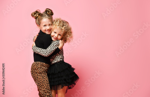 Two smiling kids girls best friends sisters in leopard print clothes pants and sweater are hugging feeling love on pink