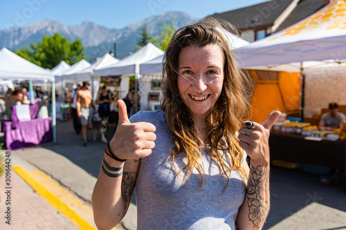 A healthy caucasian girl gives the thumbs up at local street fair for artisans and farmers, front portrait of happy shopper with market stalls behind