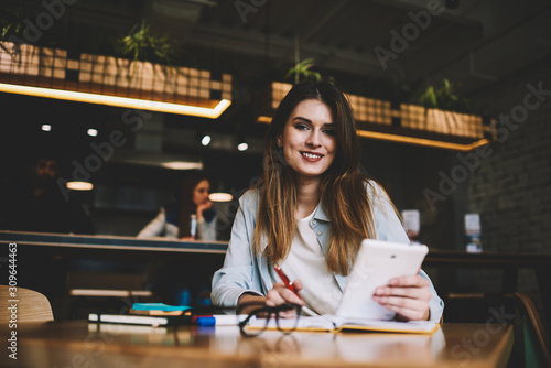 Portrait of beautiful young smiling blogger looking in camera while waiting friend at cafeteria and creating new publication, charming hipster girl holding electronic device in hand and chatting photo