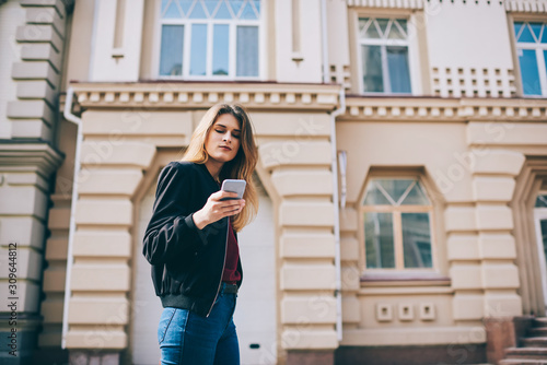 Young hipster girl resting outdoors with modern gadget connected to internet and chatting with friends, attractive thoughtful woman enjoying free time while checking notification via smartphone