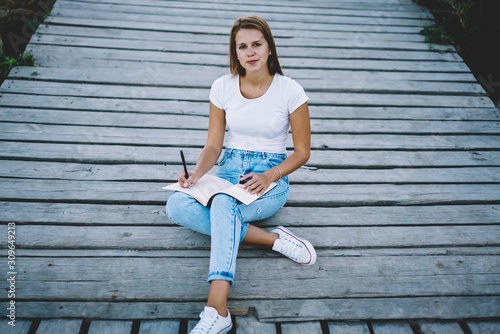 Portrait of young gorgeous female sitting outdoors with notepad , charming hipster girl in denim jeans and white t-shirt with copy space for brandname or label looking at camera resting on weekend