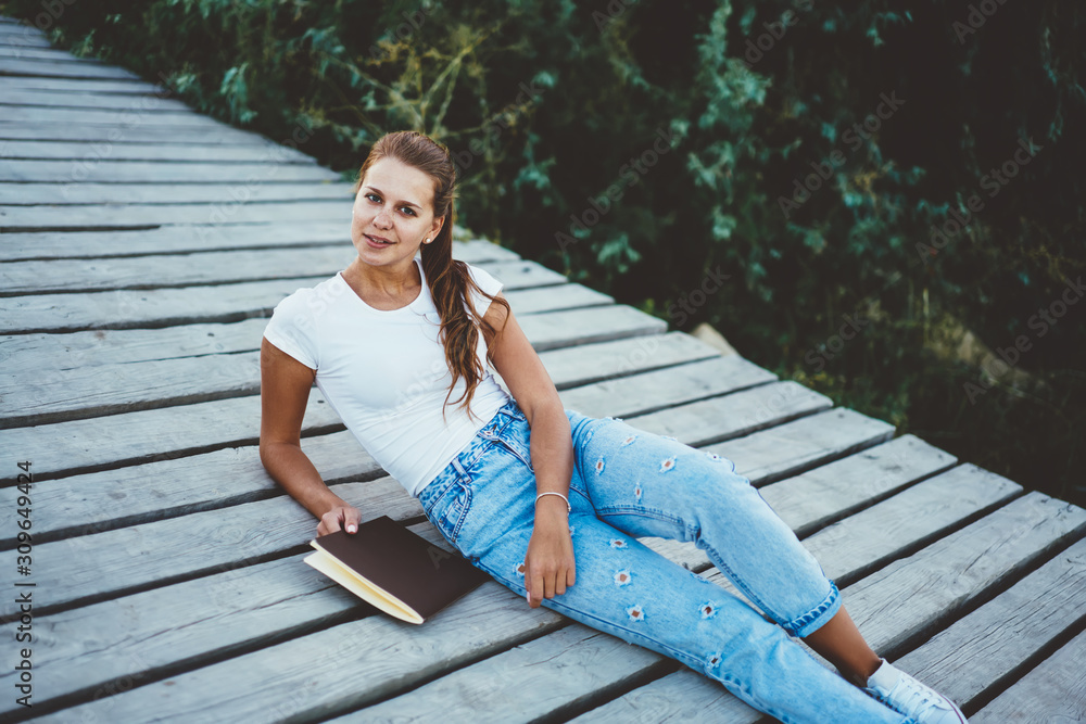 Fashionable blonde hipster girl resting at wooden pier with textbook in hand and nature on background, portrait of attractive young woman in trendy denim jeans recreating looking at camera outdoors