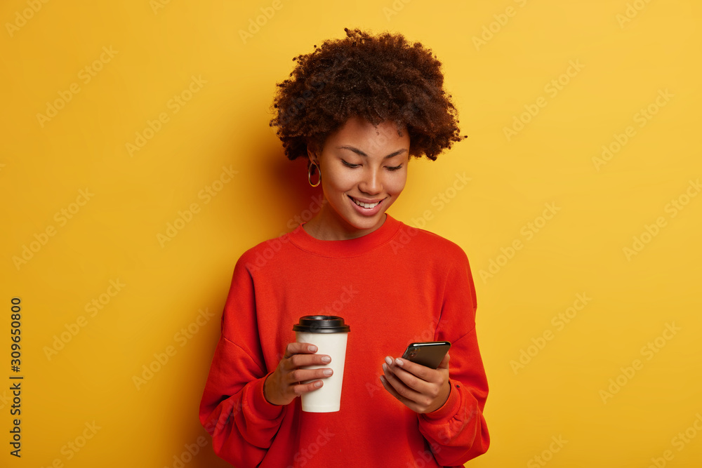 Smiling beautiful dark skinned woman holds smartphone, plays interesting game online, sends meme, drinks takeaway coffee, wears casual jumper, has Afro hairstyle, isolated on yellow background Smiling beautiful dark skinned woman holds smartphone, plays interesting game online, sends meme, drinks takeaway coffee, wears casual jumper, has Afro hairstyle, isolated on yellow background