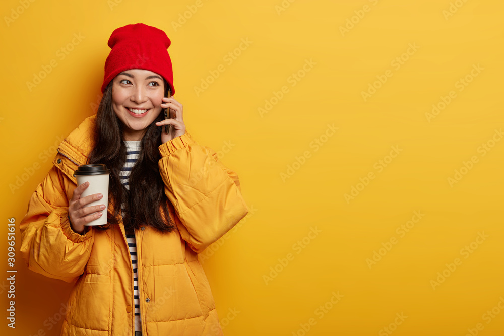 Glad millennial girl chats on mobile phone, holds paper cup of takeaway coffee, wears red hat and yellow jacket, has happy mood, shares news happened to her today, isolated on yellow background Glad millennial girl chats on mobile phone, holds paper cup of takeaway coffee, wears red hat and yellow jacket, has happy mood, shares news happened to her today, isolated on yellow background
