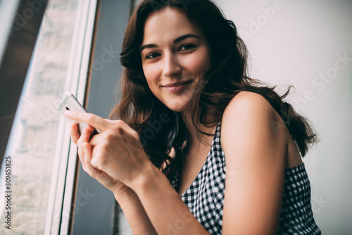 Half length portrait of positive Caucasian blogger in casual wear smiling at camera while waiting for email on cellphone using public internet connection indoors, concept of generation z woman