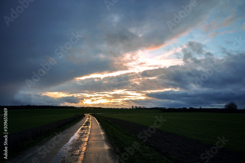 Wet reflecting country road made of concrete slabs is leading through the dark fields under a dramatic cloudy sky with evening sun, rural landscape in Mecklenburg, Germany, copy space