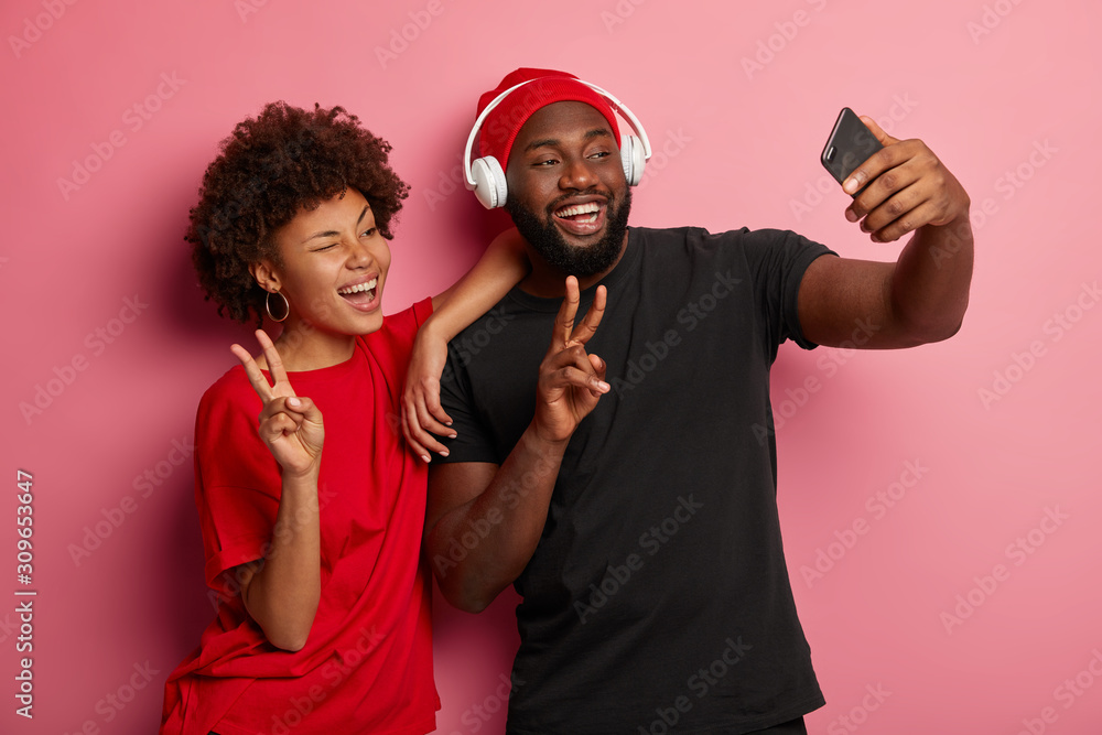 Photo of joyous ethnic female and male wink and smile broadly, make victory gesture, take selfie portrait on smartphone, dressed casually, listen music, have fun together, isolated on pink wall Photo of joyous ethnic female and male wink and smile broadly, make victory gesture, take selfie portrait on smartphone, dressed casually, listen music, have fun together, isolated on pink wall