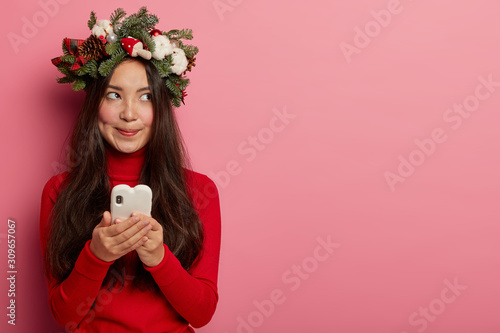 Modern technologies and communnication concept. Pensive woman with specific appearance thinks what to write in message, sends congratulation sms to friends, looks aside, isolated on pink wall photo