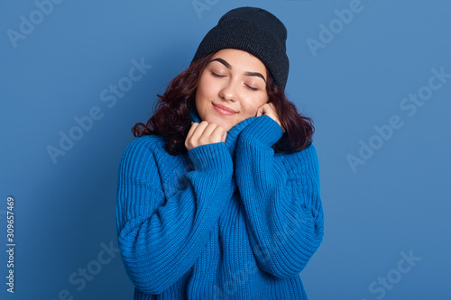Close up portrait of attractive young stylish woman wearing warm blue sweater and cap, feeling comfortable, girl likes casual style, posing isolated over studio background, standing with closed eyes.