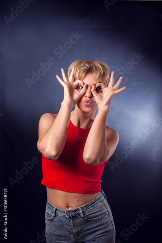 A portrait of young woman with short blond hair in red shirt showing owl or glasses symbol in front of black background. People, emotions and beauty concept