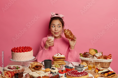 Obraz na plátne Addicted young Asian female drinks milk and eats delicious croissant, licks lips