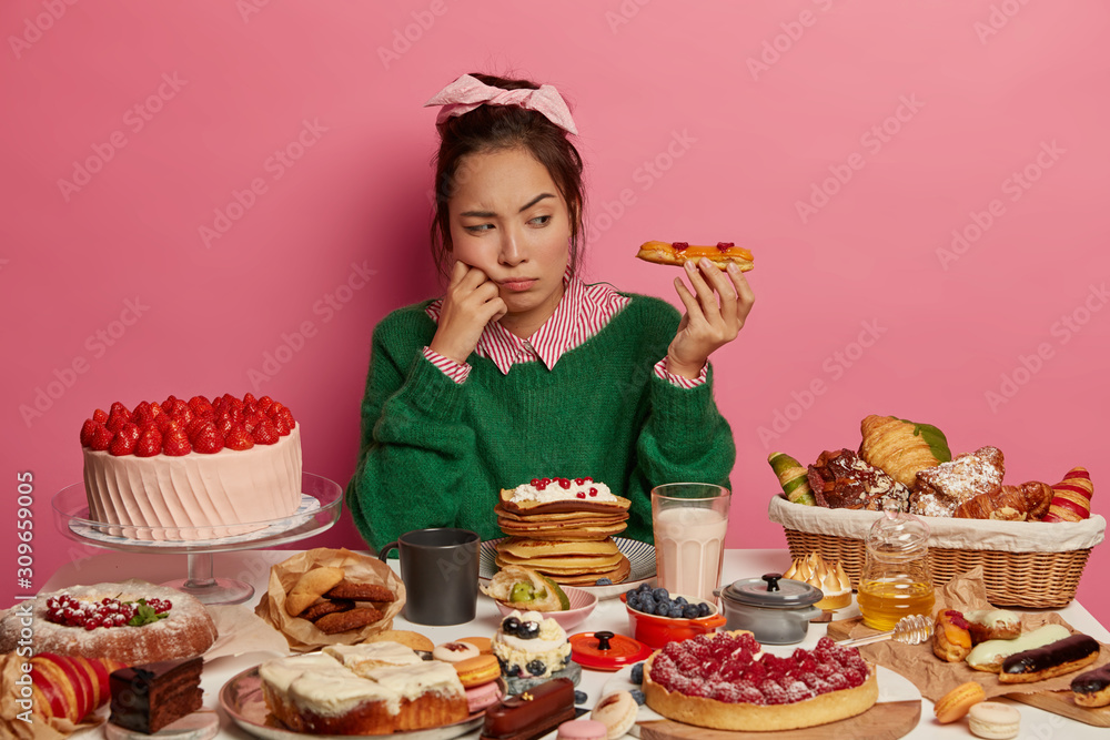 Unhappy ethnic girl looks with temptation at delicious dessert, sad as can put on weight after eating high caloried food, has variety confectionery and pasrty on table, poses in room with pink walls