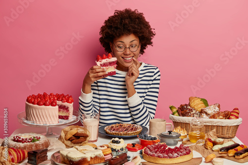 Photo of happy curly haired female holds big piece of strawberry cake, eats yummy desserts on her birthday, has sugar addiction, prepared homemade confectionery, smiles positively. Unhealthy nutrition