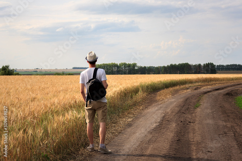 young man traveler with backpack in hat stands in field at sunset and looks away, concept of freedom of choice, future, concept of travel and adventure
