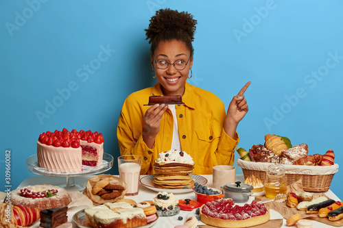 Sweet happiness. Pleased attractive dark skinned female holds tasty chocolate dessert, points over blue background, sits at table full of tasty sweet desserts waits for guests at home isolated on blue