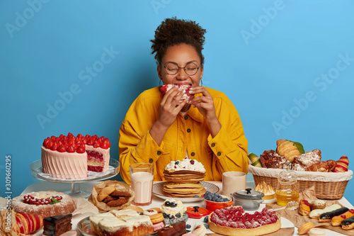 Cheat meal and gluttony concept. Ethnic curly woman eats strawberry creamy cake with much calories, has sweet lunch, tastes various desserts, leads sweet life with confectionery. Female fan of bakery photo