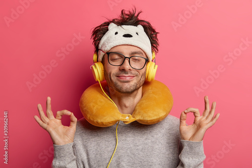 Pleased guy makes oaky gesture with both hands, meditates during listening music, has unforgettable journey, uses neck pillow for travelling on long distance, feels relaxed. People, journey, yoga, nap photo