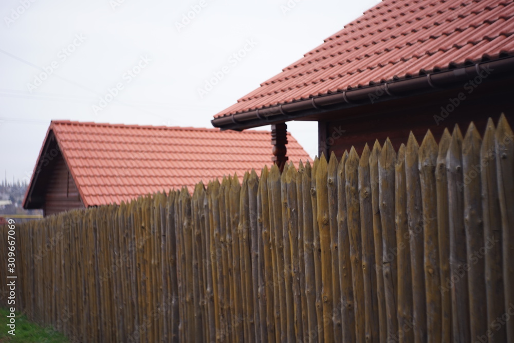 Fence made of sharp wooden stakes, wooden houses with red tiled roof and cloudy sky.