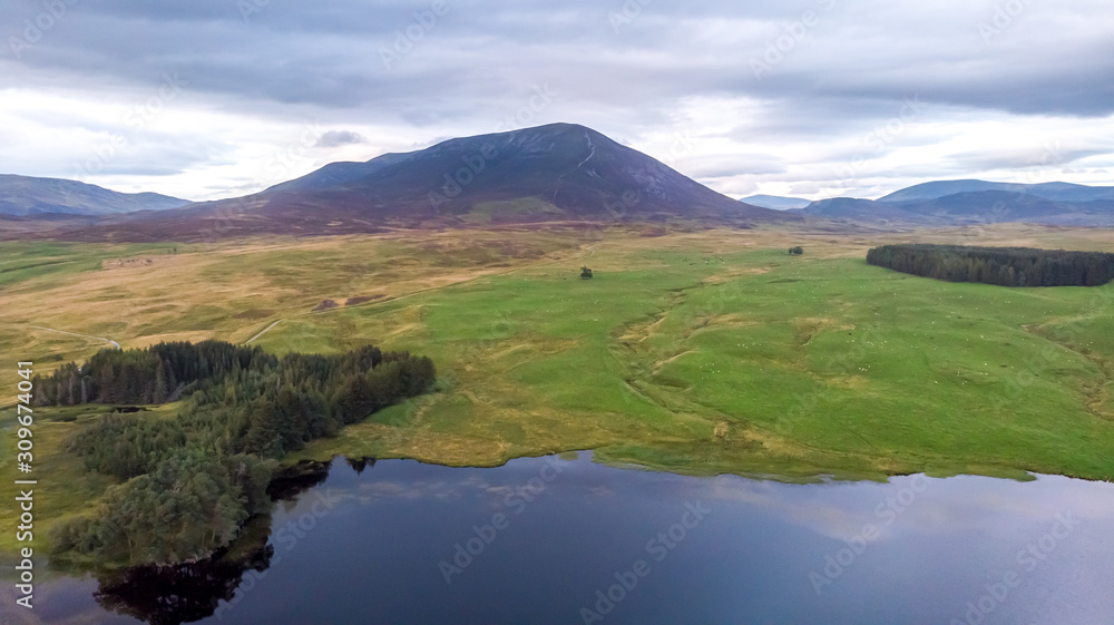 An aerial view of a majestic mountain surrounded by grassy field, trees and a lake in the foreground under a stormy sky An aerial view of a majestic mountain surrounded by grassy field, trees and a lake in the foreground under a stormy sky