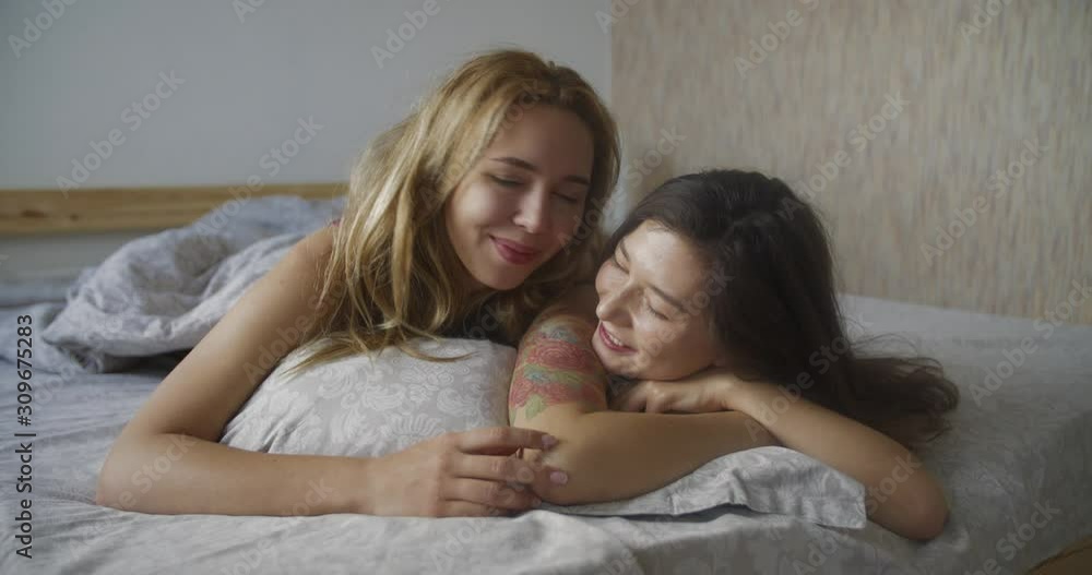Close portraits of two multinational lesbian kiss. young women lying in bed and hugging gently stroking each other's hands. LGBT, feminism Close portraits of two multinational lesbian kiss. young women lying in bed and hugging gently stroking each other's hands. LGBT, feminism