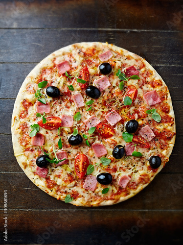 Pizza with cherry tomatoes, ham, black olives and fresh herbs. Home made food. Concept for a tasty and hearty meal. Brown wooden background. Top view. 