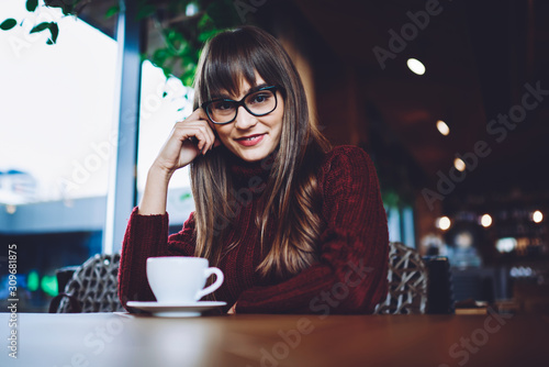 Portrait of happy charming woman in stylish spectacles sitting at cafeteria table with caffeine beverage and enjoying free time for resting, positive female teenager looking at camera and smiling