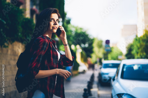 Charming positive brunette woman communicating with friends using modern smartphone during strolling at summer street.Gorgeous female person talking on mobile phone while walking at urban setting