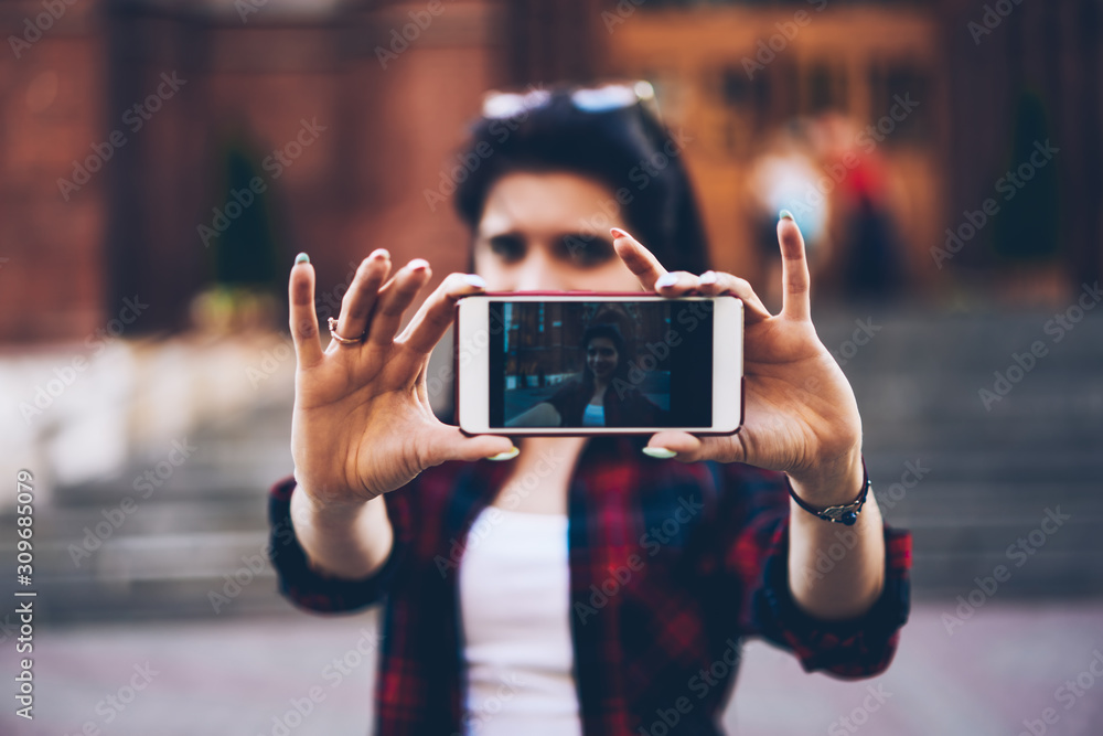 Selective focus of young female hands holding modern smartphone and taking selfie on camera with touristic place on background.Blurred image of woman dressed in trendy outfit making photo on phone