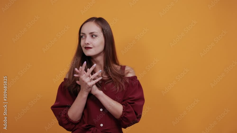 Brunette caucasian woman in maroon dress isolated on orange background in studio rubs hands, looking at camera. People sincere emotions, lifestyle concept. Brunette caucasian woman in maroon dress isolated on orange background in studio rubs hands, looking at camera. People sincere emotions, lifestyle concept.
