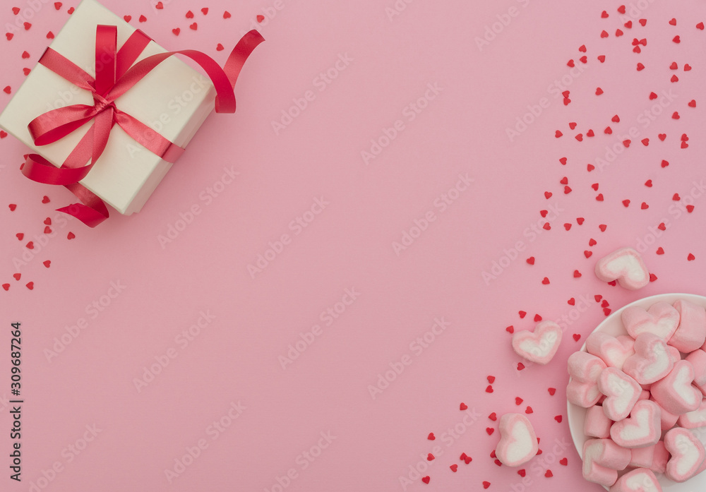 White gift box with red ribbon and heart-shaped marshmallows in white bowl on pink background with lots of small red hearts. Valentine's Day concept, greeting card. Flat lay style with copy space. White gift box with red ribbon and heart-shaped marshmallows in white bowl on pink background with lots of small red hearts. Valentine's Day concept, greeting card. Flat lay style with copy space.
