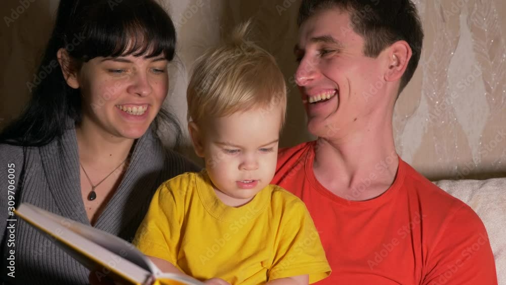 Happy Family. Young Man And Woman Read Book With Child On Sofa. Slow Motion. Evening Room Happy Family. Young Man And Woman Read Book With Child On Sofa. Slow Motion. Evening Room