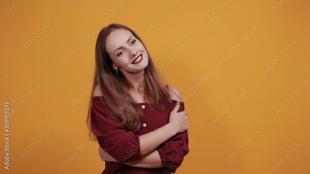 Cheerful charming lady in fashion maroon dress hugging herself, smiling isolated on orange background in studio. People sincere emotions, lifestyle concept. Cheerful charming lady in fashion maroon dress hugging herself, smiling isolated on orange background in studio. People sincere emotions, lifestyle concept.